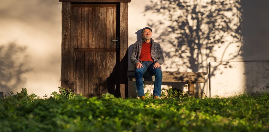 lonely senior man sitting on bench in front of old house